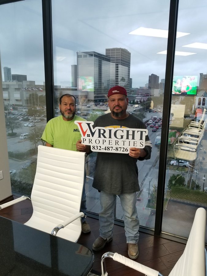 Two smiling men holding a Wright Properties sign in a Houston office with a city view